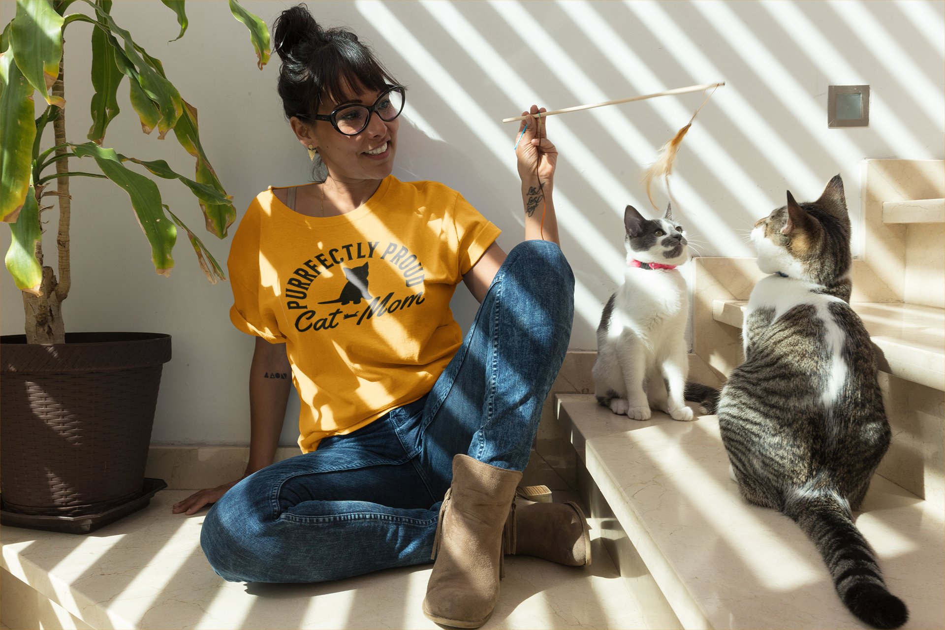 Woman wearing Purrfectly Proud Cat Mom gold long sleeve shirt playing with two cats